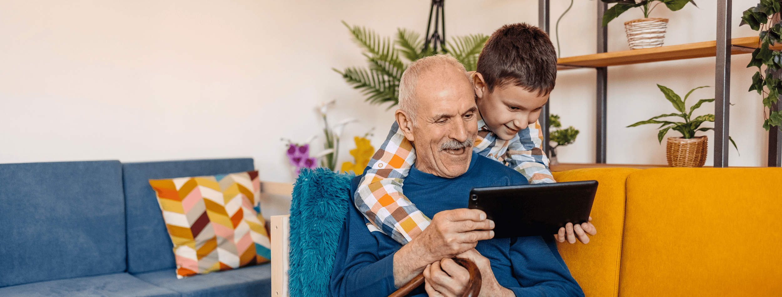 Elderly man on couch with cane and tablet in hand. Young boy hanging over shoulders helping with the tablet
